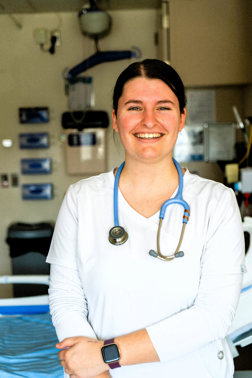 A smiling nurse with dark hair tied back stands in a hospital room. She is wearing a white scrub top, a blue stethoscope around her neck, and a smartwatch on her wrist. Medical equipment and a hospital bed are visible in the background.